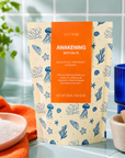 A stand-up pouch of Edit Five Awakening Bath Salts, featuring a cream background with blue sea-creature illustrations and a prominent orange label, set on a bathroom counter with towels, a blue cup, and a brush. Edit Five Awakening revitalising bath salts in sea-themed kraft packaging, designed to soothe tired muscles, refresh skin and create an invigorating minty-herbal bathing experience.