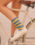 A close-up lifestyle shot of a person's legs crossed over a cream-colored bicycle, wearing blue and yellow striped socks and white sneakers. Colourful women’s wool-blend socks featuring bold blue and bright yellow stripes—a fun fashion accessory, cosy winter essential and thoughtful gift for her.