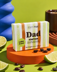 A bar of soap from Edit Five labeled 'Dad' and 'Coffee & Lime' is displayed on an orange pedestal. The soap bar has green, white, and orange vertical stripes. Luxury handmade men’s soap with exfoliating coffee and uplifting lime essential oil, creating a refreshing lather for everyday bathing, showering, skincare and grooming.