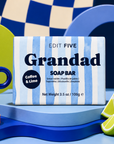 A boxed soap bar labeled 'Grandad SOAP BAR' with blue-and-white vertical stripes, placed on a blue pedestal with coffee and lime props. The visible brand is EDIT FIVE. Grandad coffee and lime soap bar, handmade in England with 100% natural essential oil, a gentle exfoliating texture and gift-ready packaging—perfect for birthdays, Father’s Day or Christmas.
