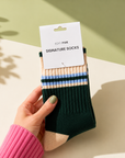 A hand holds a pair of dark green crew socks with beige and light blue stripes, still attached to a white paper hanger card that reads 'EDIT FIVE SIGNATURE SOCKS'. The background is a styled studio setup with a light beige surface and a sage green wall, featuring soft leaf shadows. Women’s green, peach and blue striped socks made from a premium soft-touch wool blend, with a warm, breathable and comfortable stretch fit for everyday wear.