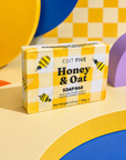 A boxed soap bar labeled 'EDIT FIVE Honey & Oat SOAP BAR' standing upright on a colorful geometric set with yellow, blue, purple, and beige shapes. Honey and oat soap bar, handmade in England using traditional methods, with a naturally comforting fragrance and rich, creamy lather for gentle everyday cleansing.