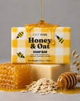 A boxed Honey & Oat soap bar sits on top of a plain soap bar, with honey dripping from above. The front box is yellow-and-white checkered with bumblebee illustrations and the text 'EDIT FIVE Honey & Oat SOAP BAR. Luxury handmade honey and oatmeal soap with a gentle cleansing formula, soft natural fragrance and creamy lather, perfect for anyone who enjoys comforting bath and body products.