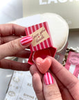 Person holding a small striped box with a heart-shaped object and 'love you loads' text.