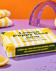A boxed lemon poppy seed soap bar sits on a lavender surface with orange and yellow props. The package is yellow and white striped with green text reading 'Lemon Poppy Seed SOAP BAR' and a tagline on the front. Edit Five lemon and poppy seed exfoliating soap bar with natural essential oil, ideal for energising morning showers, daily bathing, self-care routines and citrus lovers.