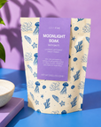 A stand-up pouch of Edit Five Moonlight Soak bath salts, featuring a lavender label and a cream background with blue sea-creature illustrations, styled on a blue and purple surface with a small bowl of white salts nearby. Moonlight Soak bath salts with Epsom and Dead Sea salts, scented with lavender, bergamot and sweet orange essential oils for calming evening baths and bedtime relaxation.