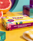 A wrapped bar of soap bar sits on a bright yellow surface with grapefruit slices, teal blocks, and a folded yellow-and-white towel around it. The soap packaging is floral and reads 'Nan SOAP BAR' and 'pink grapefruit. Edit Five pink grapefruit soap bar designed especially for Nan, featuring natural essential oils and beautiful packaging—ideal as a pampering gift, self-care present or gift-box addition.