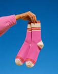 A person's hand wearing a pink knit sweater holds up a pair of pink knit socks with cream toe, heel, and cuff details, set against a solid blue sky. Colourful women’s wool-blend socks featuring vibrant pink, peach and orange stripes—a fun fashion accessory, cosy winter essential and thoughtful gift for her.