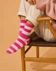 A person seated on a wooden chair wearing bright pink and white striped socks, off-white cuffed pants, and a light pink sweater against a warm beige background. Colourful women’s wool-blend socks featuring bold cherry pink and pale baby pink stripes—a fun fashion accessory, cosy winter essential and thoughtful gift for her.