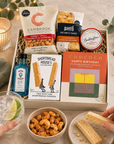 Luxury happy birthday letterbox gift hamper with birthday chocolate bar, Bombay Sapphire gin, sweet chilli peanuts and cashews, artisan shortbread, sourdough crackers and strawberry conserve in a premium Hallby gift box.