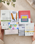 A person's hands hold a curated gift set of skincare and tea accessories against a warm beige background with soft plant and fabric props. The set includes Arthur Betay facial sheet masks, a boxed soap bar, a colorful 'Happy Birthday' card, a notebook, and scented tea lights. Hallby birthday letterbox gift for her with soap, face masks, scented tealights, warming eye mask and chocolate