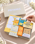 A gift box arrangement on a beige tablecloth with a hand placing a yellow card into a silver tray. The box contains tea packets, a notepad, a card, and a cookie, with a large beige gift box labeled PRIYANKA behind it. Pick me up gift for her featuring Today’s Mantra notepad, sunshine coaster and relaxing self-care treats