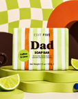 A boxed soap bar labeled 'EDIT FIVE Dad SOAP BAR' with a green, white, and orange striped package, styled on a bright green platform with lime wedges, a dark coffee mug, and abstract geometric props. Dad coffee and lime soap bar, handmade in England with natural lime essential oil, exfoliating coffee and bold gift-ready packaging—perfect for Father’s Day, birthdays or Christmas.