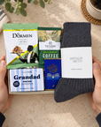 A gift box held by two hands contains curated items: a pair of dark charcoal wool socks with a white label reading Arthur Betsy, a bag of The Dormen coffee, a box of Taylor's Rich Indian Coffee, a blue box of Little & Co's, and a striped soap bar labeled Grandad. The scene is styled on a beige stone surface with a mug and plant in the background. Letterbox gift for Grandad containing Taylors coffee, The Dormen pistachios and Willie's Milk of the Gods chocolate