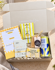 A curated gift box set displayed in an open white cardboard box with the lid propped open. The set includes a yellow-and-white notepad, a bag of Limoncello lemon balm, a packet of Arthur Betty retinol eye gel, a bag of lemon poppy seed, a small tag, and a can of Dash lemon sparkling water. Get well soon gift for her with Today’s Mantra notepad, DASH lemon water and uplifting self-care treats