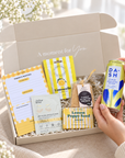 A curated gift box set displayed in an open beige box with a hand holding a can of Dash sparkling water. The box contains a yellow-and-white notepad, a lemon meringue cookie, an Arthur Bailey eye mask, and a lemon poppy seed bar, set on a beige fabric surface with soft floral background decor. Colourful gift hamper containing limoncello gummies, all-butter shortbread, lemon soap and gold under-eye mask