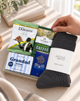 A gift set arranged on a beige fabric surface, featuring a dark charcoal wool sock with a white Arthur Betsy band, a soap bar, a coffee bag, and a snack bag, with a hand holding the sock. A cup of coffee, books, and greenery are visible in the background. Hallby Grandad letterbox gift with wool socks, coffee, pistachios, chocolate and Grandad soap
