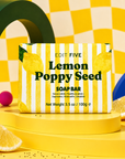 A lemon poppy seed soap bar by Edit Five, presented in its yellow and white striped packaging on a yellow pedestal, surrounded by decorative yellow and green props. Lemon poppy seed soap bar, handmade in England with 100% natural lemon essential oil, gently exfoliating poppy seeds and bright gift-ready packaging for refreshing daily skincare.