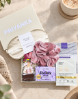 A curated gift set from Arthur Betsey and Priyanka, beautifully arranged in a flat lay. The set includes a cream satin sleep mask, a dusty pink silk scrunchie, a purple 'Relax' hair mask, an eye mask packet, and a chocolate bar, all presented in a cream-colored gift box with the brand names clearly visible. Hallby letterbox gift for her with Relax soap, warming eye mask, satin eye mask and hot chocolate