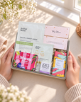 A gift box of skincare and tea products held by two hands on a cozy windowsill. The box contains Arthur Betsy facial sheet masks, a pink 'My Mum' gift card, a bag of Joe's Tea Teacake, and a colorful box of Oreo chocolate. Mother’s Day letterbox gift with self-care treats, Joe’s Tea, chocolate and a meaningful string bracelet