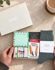 A curated gift set for Dad laid out on a light stone surface, held by two hands. The set includes a cream gift box labeled DAD, a green patterned note card, a black and red Taylors English Breakfast Tea packet, a Shortbread House of Edinburgh biscuit packet, a packet of dad-themed shortbread papers, and a pair of dark green Arthur Betsy wool socks with a white label. Hallby letterbox gift for Dad with Twilight wool socks, Dad soap, shortbread, English Breakfast tea and a Better Write This Down memo pad