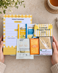 A styled flat-lay showing a curated gift set held by two hands. The set includes a yellow and white planner pad titled 'TODAY'S MANTRA', Twinin tea packets (Defence, Detox, and Lemon Poppy Seed), an Arthur Betty eye mask packet, a RAW HALO supplement box, and a Yorkshire Flapjack cookie. Hallby sunshine letterbox gift for her with positive notepad, bath salts, face mask, soap and chocolate