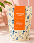 A stand-up pouch of Edit Five Awakening Bath Salts, featuring a cream-colored background with blue sea-themed illustrations and a prominent orange label, set on a tiled surface with a pink wall and a green plant in the background. Awakening bath salts with Epsom and Dead Sea salts, scented with eucalyptus, peppermint and rosemary essential oils for an energising, refreshing and uplifting bath.