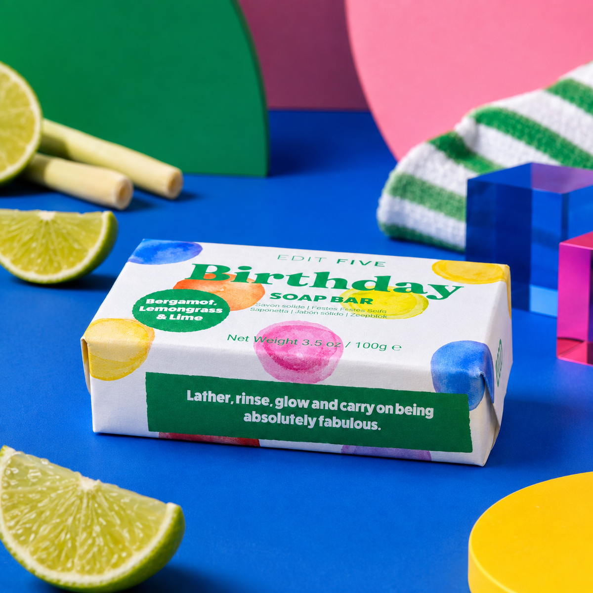 A boxed soap bar labeled 'Birthday' by Edit Five, styled on a bright blue surface with lime wedges, colorful geometric shapes, and a striped towel in the background. Edit Five natural essential-oil birthday soap bar in playful gift-ready packaging, ideal as a birthday gift for her, birthday gift for him, stocking filler or pamper hamper addition.