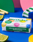 A boxed soap bar labeled 'Birthday' by Edit Five, styled on a bright blue surface with lime wedges, colorful geometric shapes, and a striped towel in the background. Edit Five natural essential-oil birthday soap bar in playful gift-ready packaging, ideal as a birthday gift for her, birthday gift for him, stocking filler or pamper hamper addition.