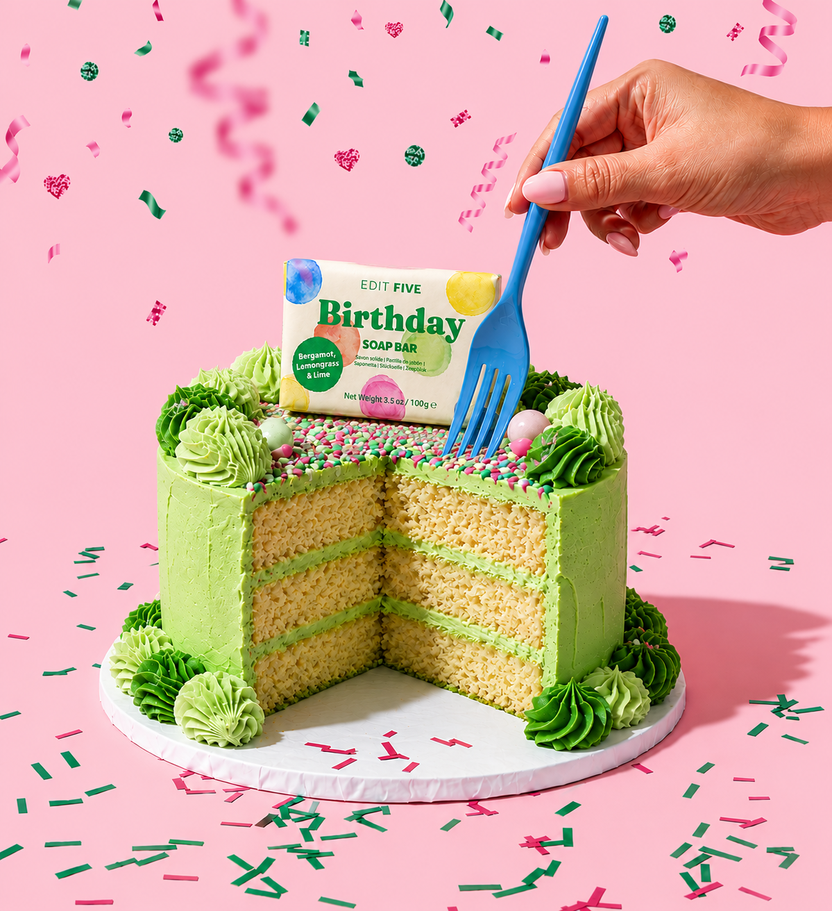A green frosted birthday cake with a slice cut out, topped with a bar of 'Edit Five' Birthday Soap Bar, and a hand holding a blue plastic fork against a pink confetti background. Luxury handmade citrus soap featuring bergamot, lemongrass and lime essential oils, a rich glycerin lather and bright confetti artwork for birthday celebrations.