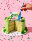 A green frosted birthday cake with a slice cut out, topped with a bar of 'Edit Five' Birthday Soap Bar, and a hand holding a blue plastic fork against a pink confetti background. Luxury handmade citrus soap featuring bergamot, lemongrass and lime essential oils, a rich glycerin lather and bright confetti artwork for birthday celebrations.