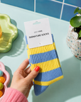A hand holds a pair of blue and yellow striped socks on a white paper hanger card, set against a styled tabletop with a plant, colorful candies, and a glass on a tray. Women’s blue and yellow striped socks made from a soft, warm and breathable wool-blend fabric, with a comfortable stretch fit for cosy everyday wear.