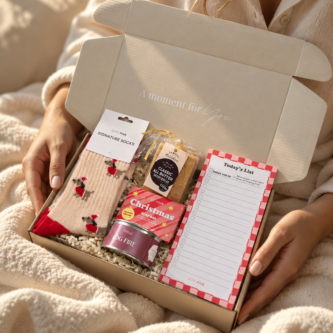 A cozy gift box held by hands wrapped in a cream blanket. The open box contains a Christmas-themed assortment including a tin of Peppa Pig Christmas treats, a packet of Christmas cookies, a notepad, and a pair of red-and-white patterned socks. Hallby Christmas gift box with Dachshund socks, list pad, Log Fire candle, all-butter shortbread and Christmas soap