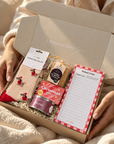 A cozy gift box held by hands wrapped in a cream blanket. The open box contains a Christmas-themed assortment including a tin of Peppa Pig Christmas treats, a packet of Christmas cookies, a notepad, and a pair of red-and-white patterned socks. Hallby Christmas gift box with Dachshund socks, list pad, Log Fire candle, all-butter shortbread and Christmas soap