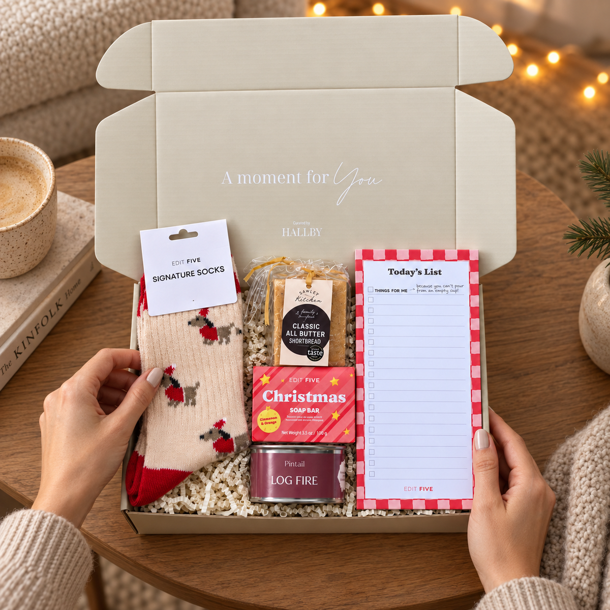 A curated gift box opened by two hands, filled with cozy holiday items including red-and-white patterned socks, a tin of Christmas soap, a log fire candle, a list pad, and a packaged chocolate. The box is set on a wooden table with warm string lights and a mug in the background. Christmas wellbeing gift in a Hallby mailer box with cosy socks, candle, stationery, shortbread and festive soap
