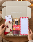 A curated gift box opened by two hands, filled with cozy holiday items including red-and-white patterned socks, a tin of Christmas soap, a log fire candle, a list pad, and a packaged chocolate. The box is set on a wooden table with warm string lights and a mug in the background. Christmas wellbeing gift in a Hallby mailer box with cosy socks, candle, stationery, shortbread and festive soap
