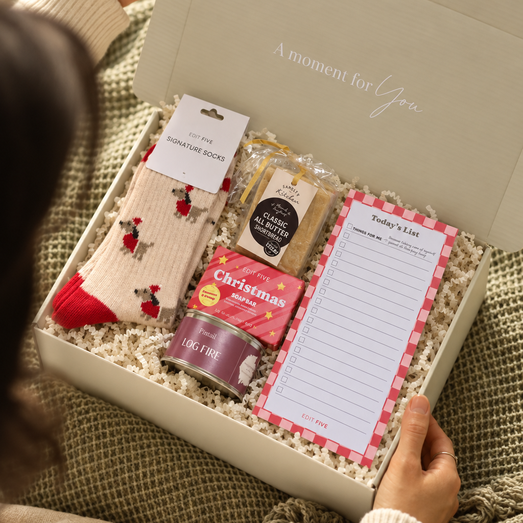 A cozy gift box set being held open by a person sitting on a knitted blanket. The white box contains a pair of cream socks with red holiday motifs, a tin of Christmas biscuits, a packet of tea, and a lined writing pad on shredded paper. Curated Christmas gift box for her featuring Edit Five Dachshund socks, Christmas soap and a Pintail candle
