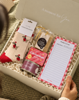 A cozy gift box set being held open by a person sitting on a knitted blanket. The white box contains a pair of cream socks with red holiday motifs, a tin of Christmas biscuits, a packet of tea, and a lined writing pad on shredded paper. Curated Christmas gift box for her featuring Edit Five Dachshund socks, Christmas soap and a Pintail candle
