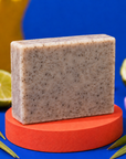 A rectangular bar of speckled soap in a light beige-gray tone sits on a round orange pedestal against a bright blue background, with lime slices and green leaves used as styled props. Handmade 100g artisan soap for Dad, combining invigorating coffee with fresh lime essential oil for an energising fragrance and gently exfoliating everyday cleanse.
