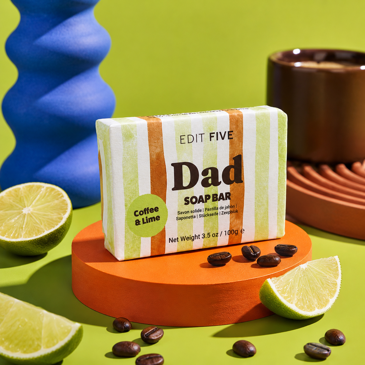 A bar of soap from Edit Five labeled 'Dad' and 'Coffee &amp; Lime' is displayed on an orange pedestal. The soap bar has green, white, and orange vertical stripes. Luxury handmade men’s soap with exfoliating coffee and uplifting lime essential oil, creating a refreshing lather for everyday bathing, showering, skincare and grooming.
