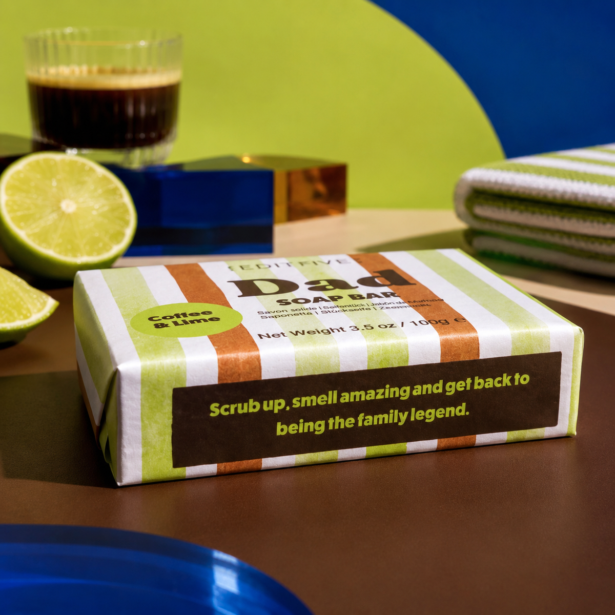 A boxed soap bar labeled 'Dad SOAP BAR' with green, white, and orange stripes sits on a tabletop. The package has a dark brown label reading 'Coffee &amp; Lime' and a slogan. Edit Five coffee and lime soap bar designed especially for dads, featuring 100% natural essential oils and colourful packaging—a practical Father’s Day gift or birthday present.