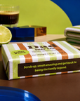 A boxed soap bar labeled 'Dad SOAP BAR' with green, white, and orange stripes sits on a tabletop. The package has a dark brown label reading 'Coffee & Lime' and a slogan. Edit Five coffee and lime soap bar designed especially for dads, featuring 100% natural essential oils and colourful packaging—a practical Father’s Day gift or birthday present.