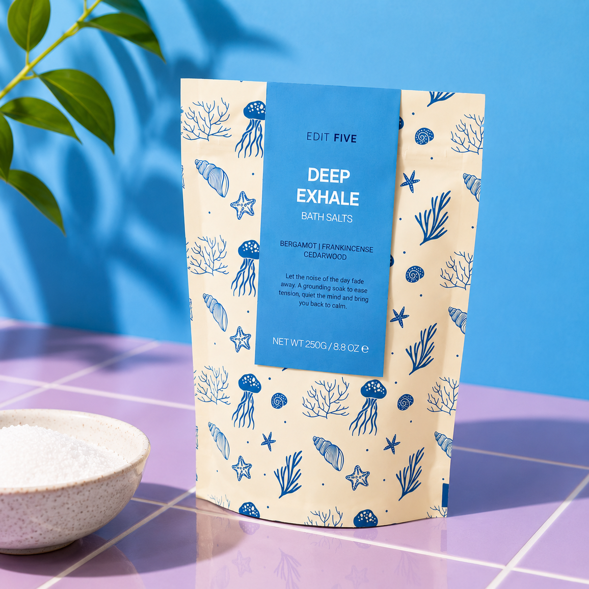 A stand-up pouch of Edit Five Deep Exhale Bath Salts stands on a purple tiled surface against a bright blue background. The packaging is cream with blue ocean-themed illustrations including starfish, sea urchins, and coral, with a blue central label. Deep Exhale bath salts with Epsom and Dead Sea salts, scented with bergamot, frankincense and cedarwood essential oils for a calming, grounding evening bath.