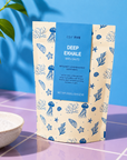A stand-up pouch of Edit Five Deep Exhale Bath Salts stands on a purple tiled surface against a bright blue background. The packaging is cream with blue ocean-themed illustrations including starfish, sea urchins, and coral, with a blue central label. Deep Exhale bath salts with Epsom and Dead Sea salts, scented with bergamot, frankincense and cedarwood essential oils for a calming, grounding evening bath.
