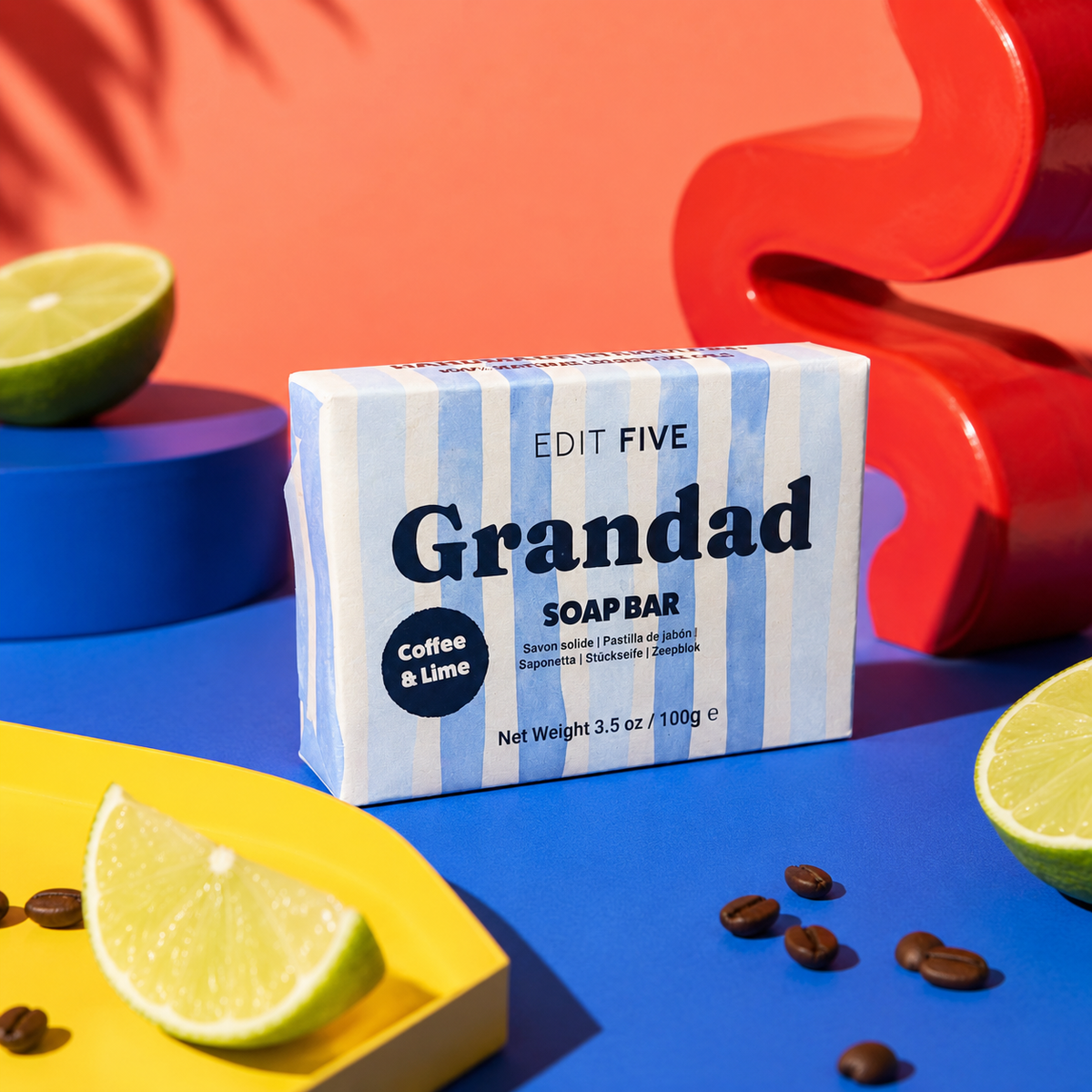 A boxed soap bar labeled 'Grandad SOAP BAR' with 'Coffee &amp; Lime' sits on a bright blue surface, surrounded by lime wedges, coffee beans, and bold decorative props in orange, red, and yellow. The soap box is the main product, and no person or real environment is present, so this is Product/Packaging rather than Lifestyle. Luxury handmade soap for Grandad with exfoliating coffee and uplifting lime essential oil, creating a fresh, invigorating lather for everyday bathing, showering and men’s self-care.