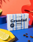 A boxed soap bar labeled 'Grandad SOAP BAR' with 'Coffee & Lime' sits on a bright blue surface, surrounded by lime wedges, coffee beans, and bold decorative props in orange, red, and yellow. The soap box is the main product, and no person or real environment is present, so this is Product/Packaging rather than Lifestyle. Luxury handmade soap for Grandad with exfoliating coffee and uplifting lime essential oil, creating a fresh, invigorating lather for everyday bathing, showering and men’s self-care.