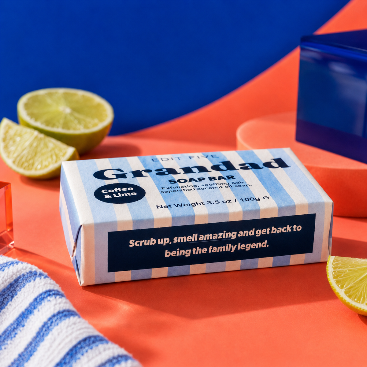 A boxed soap bar labeled 'Grandad' with 'Coffee &amp; Lime' scent, shown on a bright orange surface with sliced lime and a blue-and-white striped towel nearby. The product box is the main subject, and no person or animal is present. Edit Five coffee and lime soap bar designed especially for Grandad, featuring natural essential oils and colourful packaging—a practical birthday gift, Christmas present or Father’s Day gift.