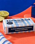 A boxed soap bar labeled 'Grandad' with 'Coffee & Lime' scent, shown on a bright orange surface with sliced lime and a blue-and-white striped towel nearby. The product box is the main subject, and no person or animal is present. Edit Five coffee and lime soap bar designed especially for Grandad, featuring natural essential oils and colourful packaging—a practical birthday gift, Christmas present or Father’s Day gift.