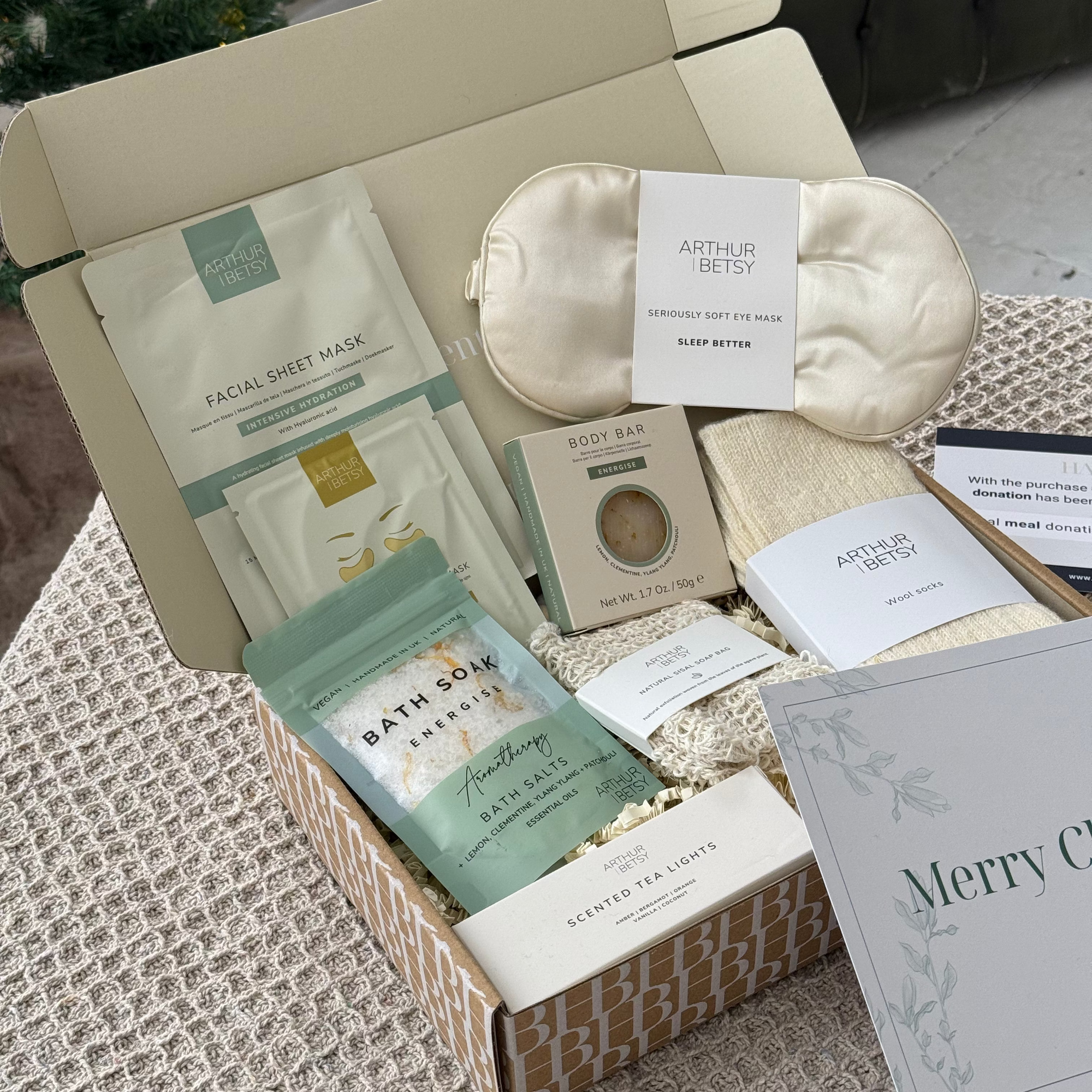 Opened gift box with self-care items including a facial mask, bath soap, and eye mask on a textured surface.