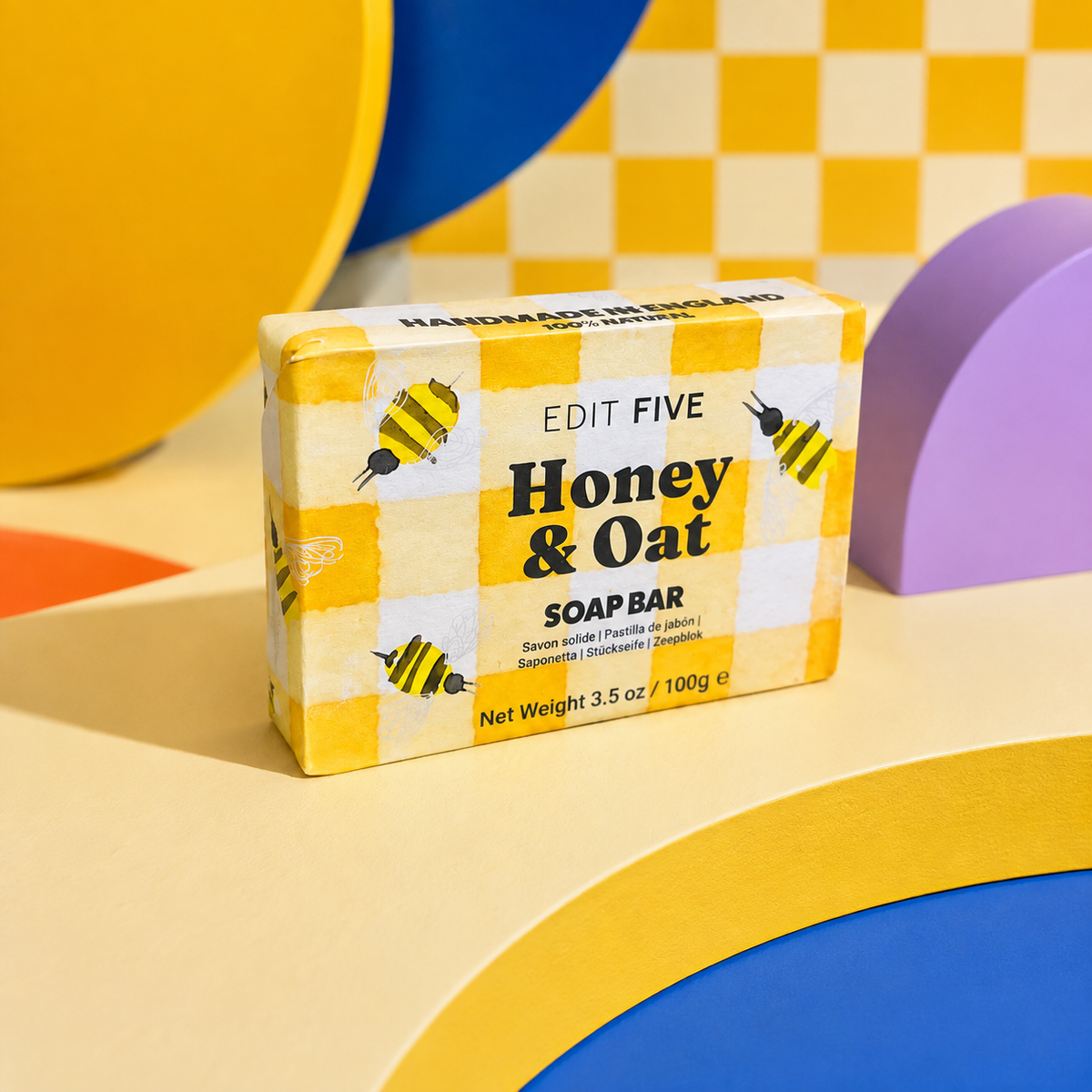 A boxed soap bar labeled 'EDIT FIVE Honey &amp; Oat SOAP BAR' standing upright on a colorful geometric set with yellow, blue, purple, and beige shapes. Honey and oat soap bar, handmade in England using traditional methods, with a naturally comforting fragrance and rich, creamy lather for gentle everyday cleansing.
